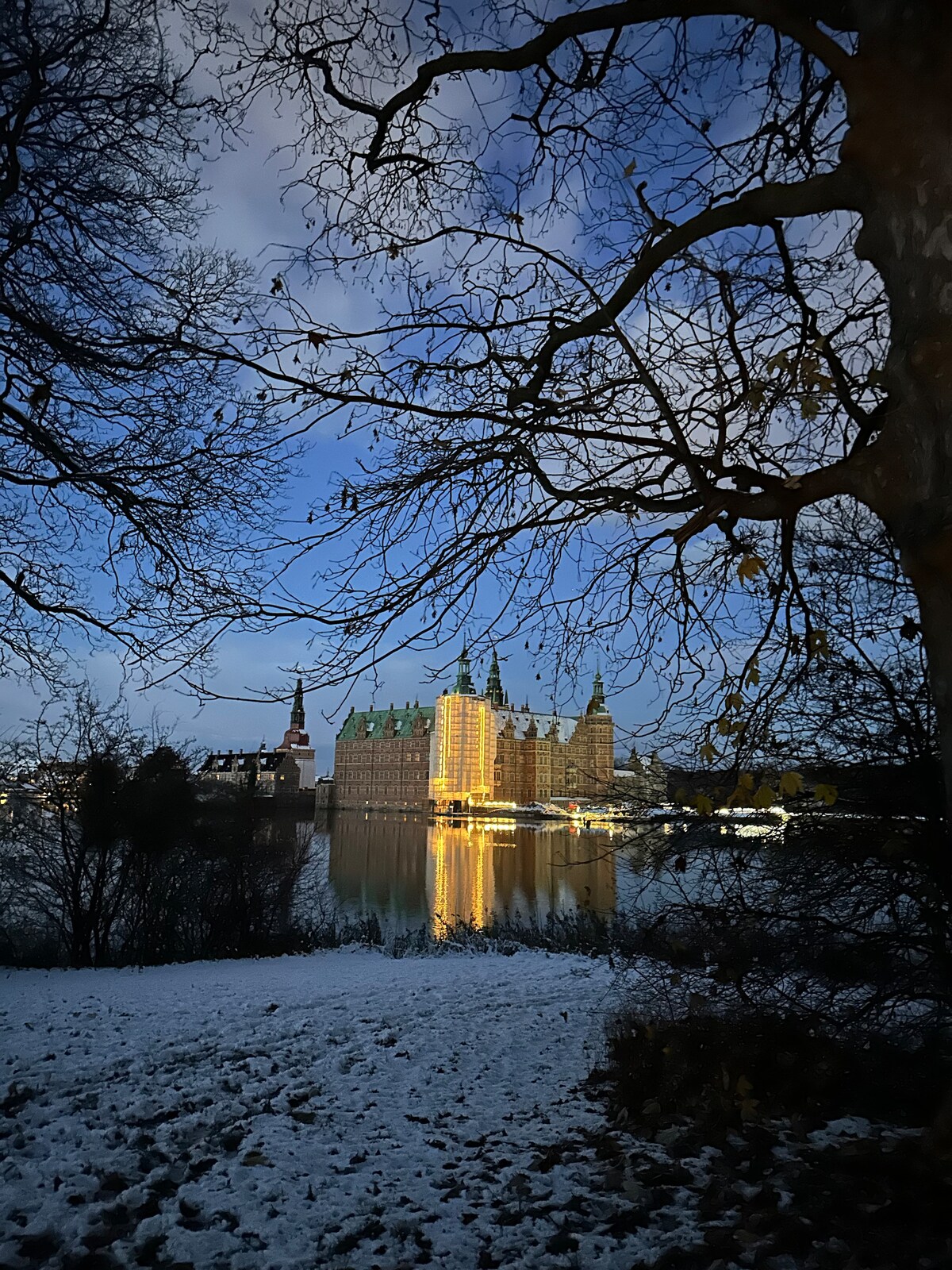A glimpse of Frederiksborg Castle is captured through bare branches, with soft lighting reflecting off the water. The serene winter landscape is adorned with a light dusting of snow, enhancing the tranquil atmosphere of the scene.
