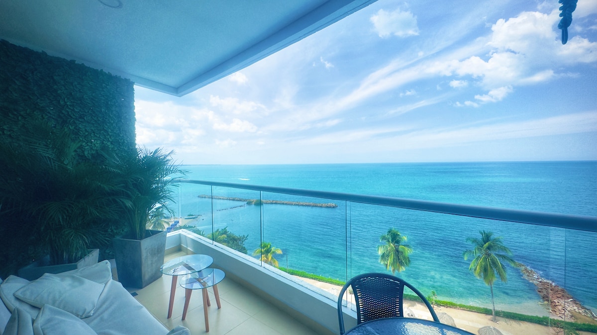 An outdoor balcony area offers stunning views of the ocean, with bright blue waters meeting the sky. A comfortable seating arrangement features a plush sofa and two small tables, surrounded by greenery and a palm tree, enhancing the tranquil atmosphere.