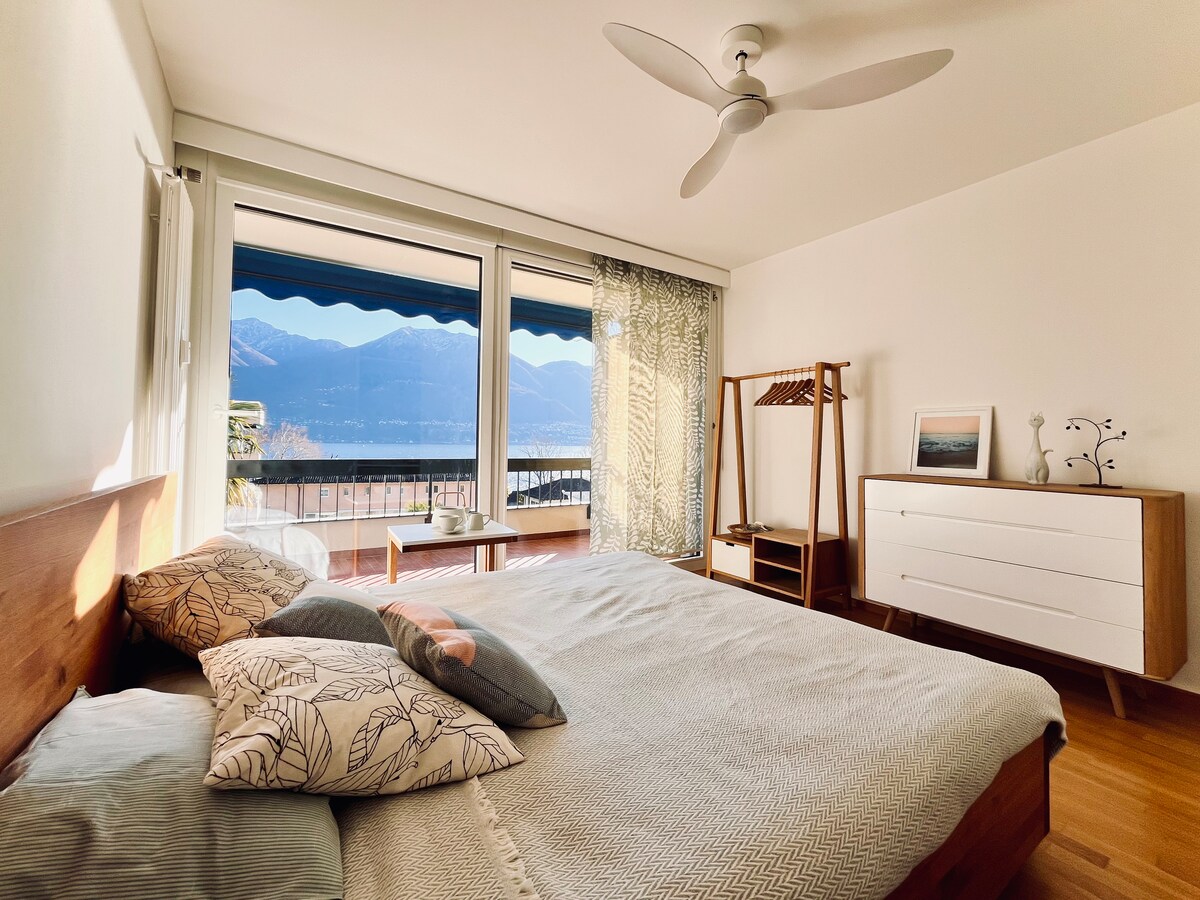 The bedroom is designed with a spacious bed adorned with decorative pillows. A large window offers a panoramic view of the lake and mountains, while a ceiling fan provides airflow. Nearby, a modern dresser and a clothing rack enhance functionality.