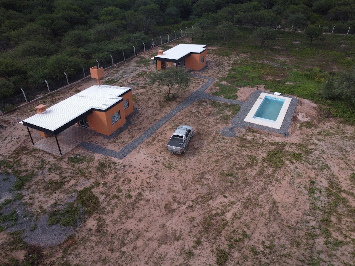 An aerial view showcases two separate structures with flat roofs, surrounded by natural landscaping. A car is parked on a gravel path that leads to a private pool, highlighting open space and the tranquility of the surrounding environment.