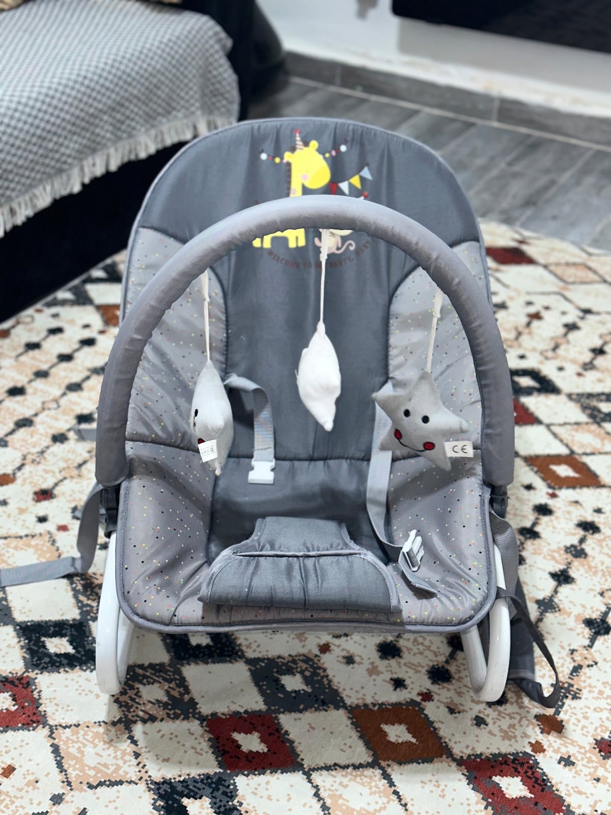 A gray baby bouncer is positioned on a patterned area rug. The seat features playful toys hanging from the arch, including a soft gray cat and a white plush object. The background shows a cozy sofa and a textured throw.