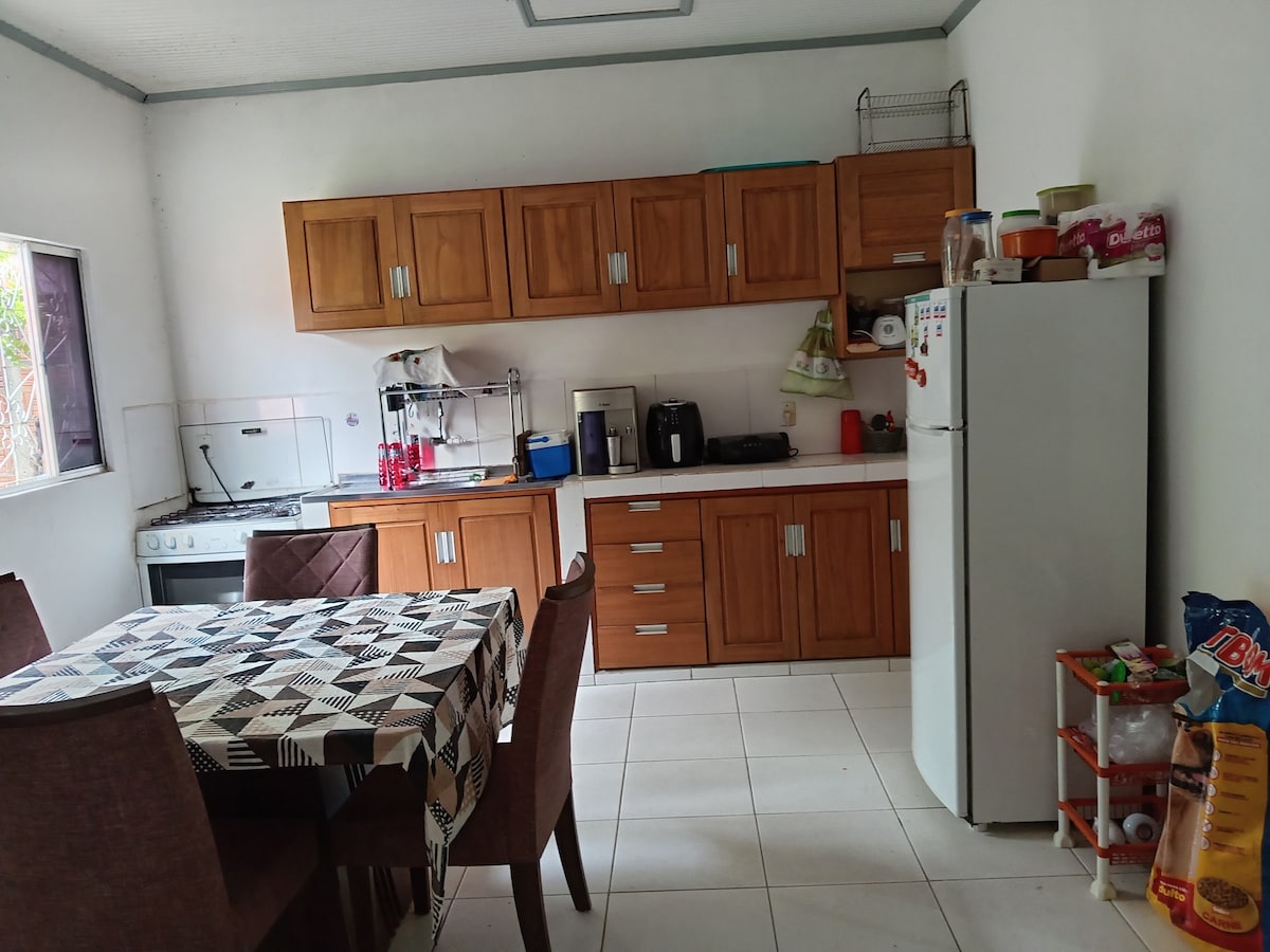 A kitchen area is presented, featuring wooden cabinets and a spacious countertop. Essential appliances, including a refrigerator, coffee maker, and stove, are visible. A dining table set for four is placed in the center, and a variety of kitchen items are neatly arranged for convenience.