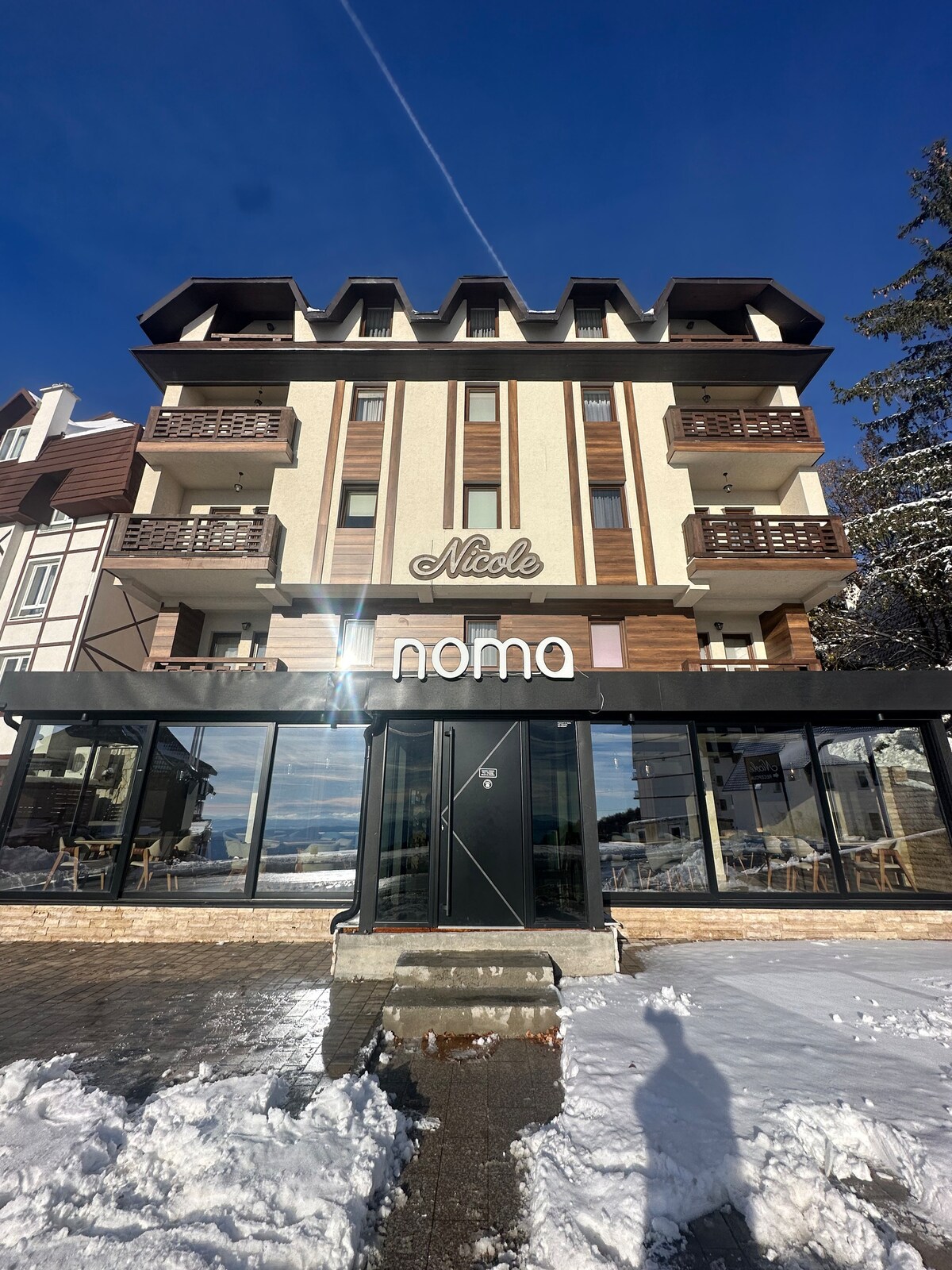 The exterior of a multi-story hotel building is presented, featuring a blend of dark wood and light stone. Large glass windows reflect sunlight, providing views of the surrounding area. A sign reading 'nomo' is prominently displayed near the entrance, with snow covering the ground.