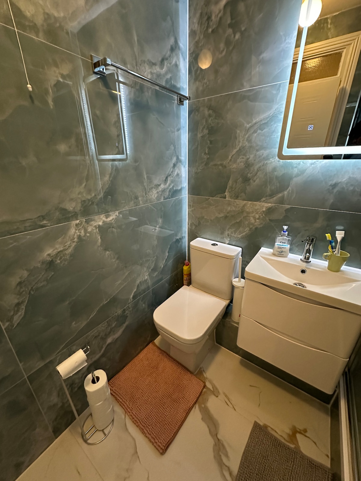 A modern bathroom features sleek marbled walls complemented by a compact vanity and a contemporary toilet. A mirror with integrated lighting is mounted above the sink, enhancing visibility. A small rug is placed on the floor, with essential toiletries neatly arranged on the vanity.