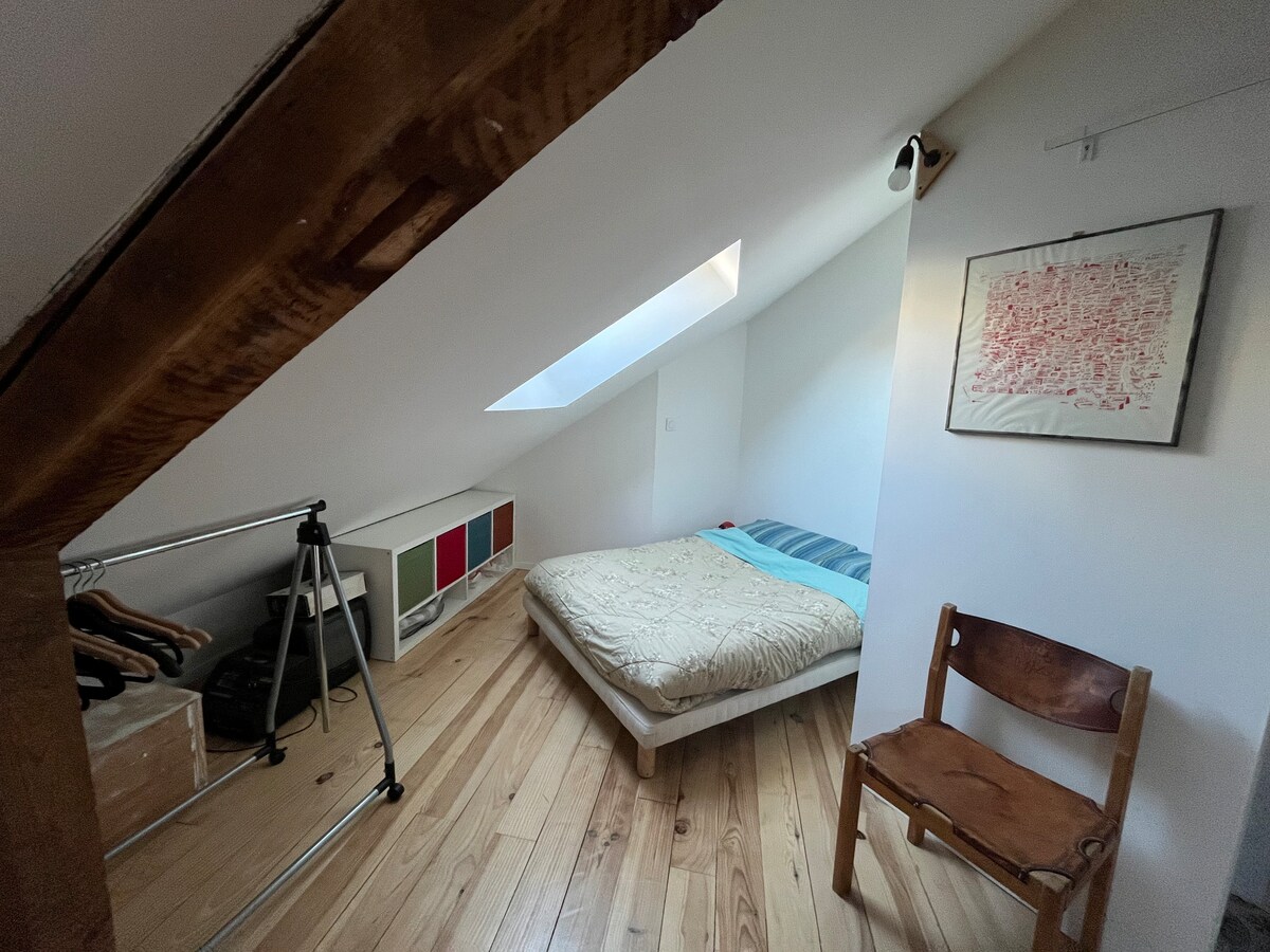 A cozy sleeping area is highlighted by a spacious bed with a patterned coverlet. Natural light filters through a skylight, illuminating the warm wooden floor. A simple wooden chair and a clothing rack are visible, alongside stylish storage units and a wall art piece.