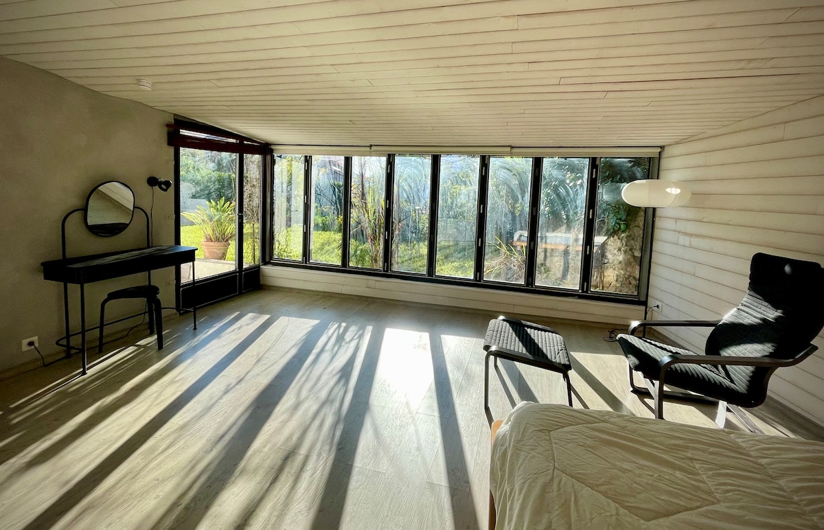 A spacious room is illuminated by natural light streaming through large glass doors. The space includes a black desk and chair, a modern lounge chair, and a comfortable bed. Views of the garden create a serene backdrop, enhancing the sense of tranquility.