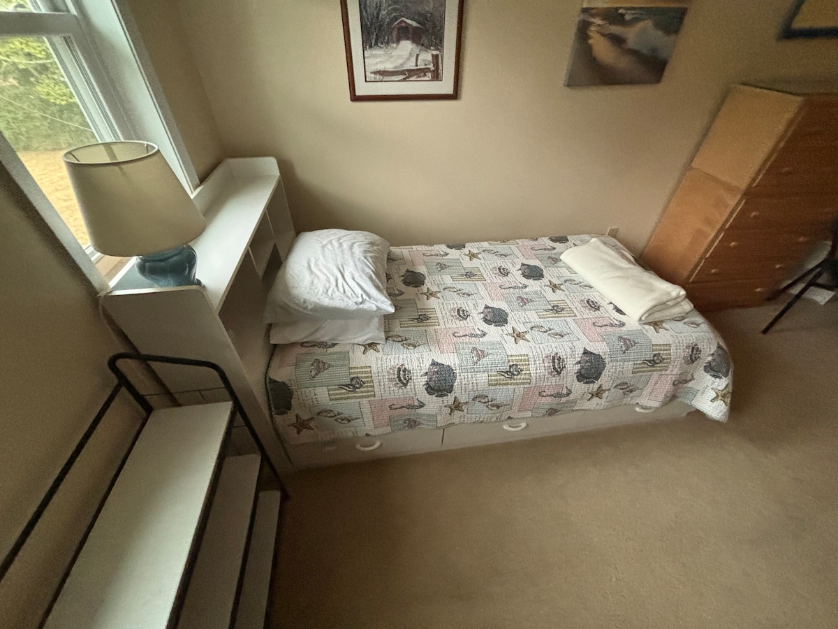 A cozy bedroom features a twin bed with patterned bedding, complemented by a light-colored quilt. A bedside lamp provides soft lighting. A wooden dresser and a small shelf are positioned along the walls, while a window invites natural light into the space.