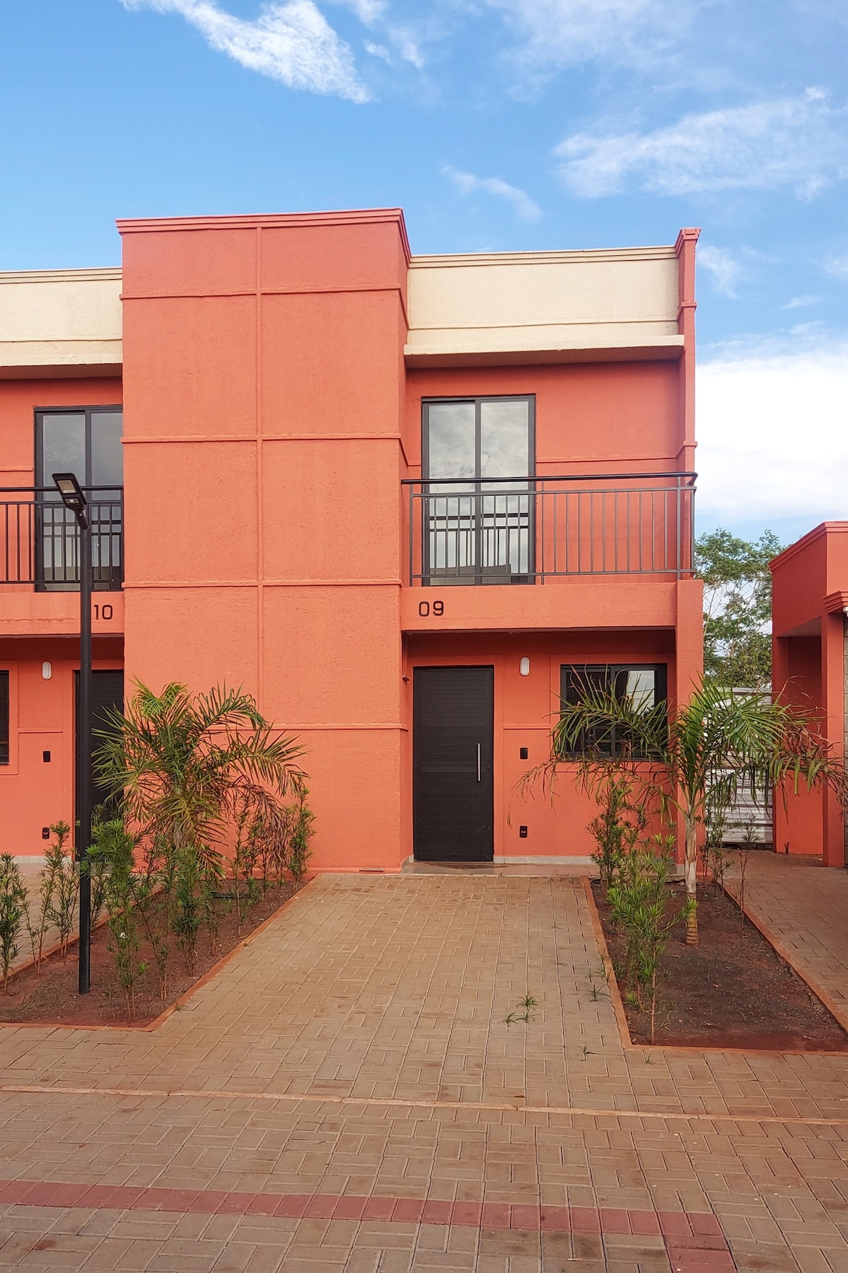 A duplex structure features a vibrant coral exterior, characterized by large windows and a balcony. Adjacent greenery and freshly manicured landscaping enhance the entrance, which is set on a paved pathway, leading to the main door.