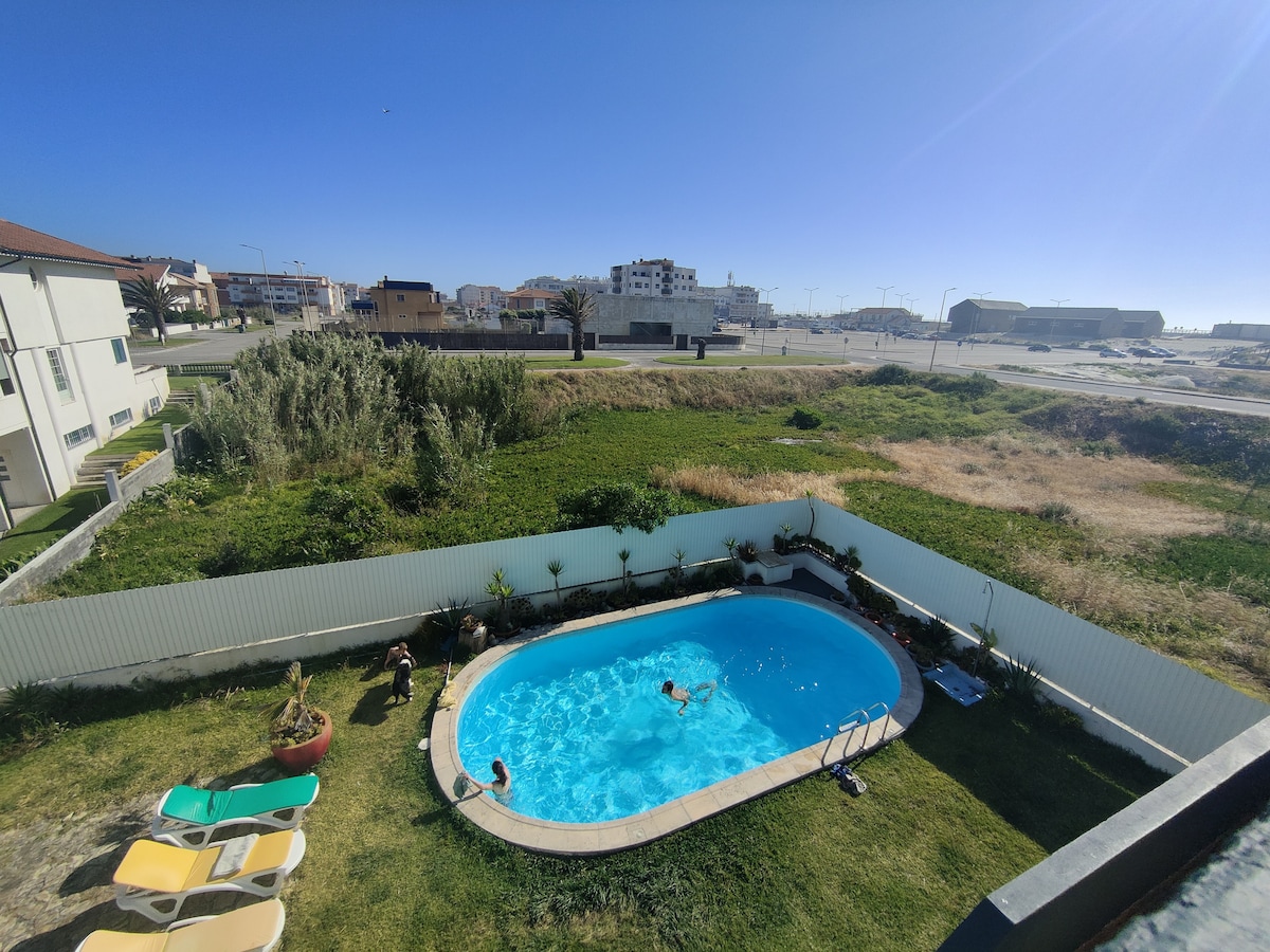 An outdoor view features a clear blue swimming pool surrounded by green grass. Sun loungers are positioned nearby, and children can be seen playing in the water. Fencing and greenery define the pool area, while distant buildings and a road are observable in the background.
