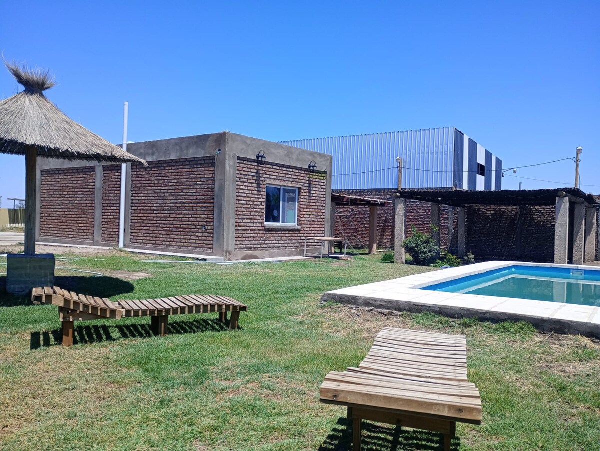 A sunlit outdoor area features a clear pool bordered by a stone deck. Nearby, wooden lounge chairs are arranged on the lush green grass. A thatched roof gazebo provides shade, while a rustic brick building is visible in the background.