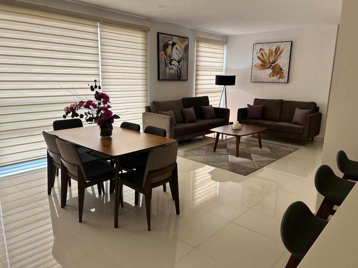A spacious living area features a modern dining table surrounded by chairs, complemented by a cozy seating arrangement. Soft lighting enhances the atmosphere, while large windows with blinds allow natural light to fill the room. Art pieces decorate the walls, adding sophistication.