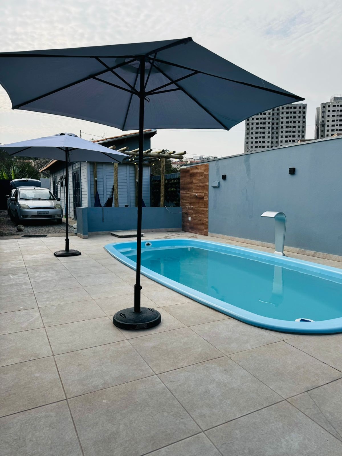 A refreshing pool is positioned in a paved area, complemented by two strategically placed umbrellas providing shade. The light blue water reflects the sky, while a wall in the background adds privacy to the space. Parking is visible in the background behind the pool.