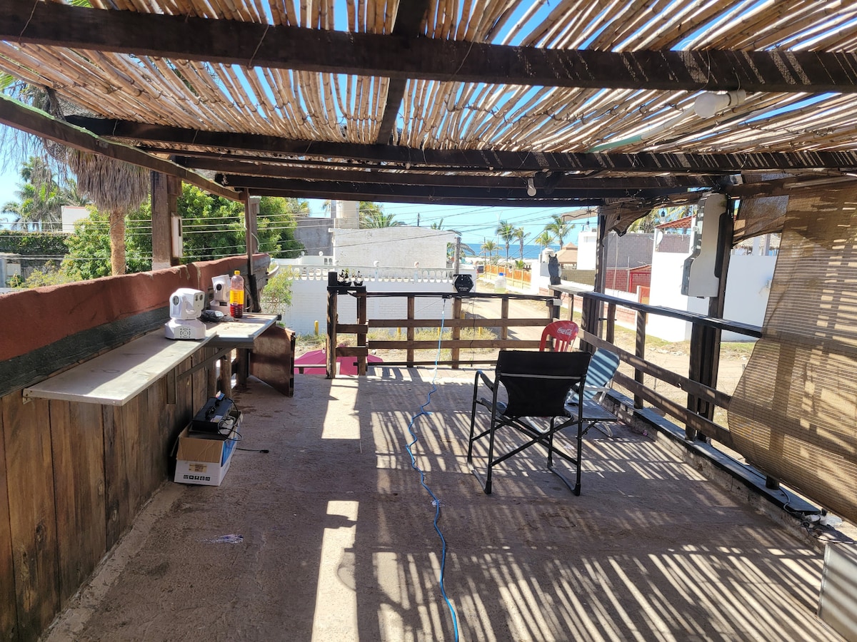 An open terrace is presented, featuring wooden beams and a thatched roof providing shade. A simple table and a chair invite relaxation, while the nearby railing offers a view of the surroundings. A soft light is seen filtering through the overhead structure.