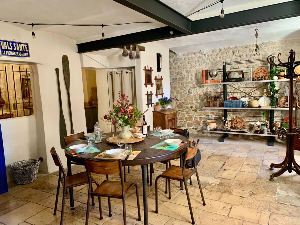 An inviting dining area features a circular wooden table set for six, surrounded by wooden chairs. Natural light illuminates the stone walls showcasing decorative items and rustic decor. A side shelf displays handcrafted pieces and plants, contributing to the authentic atmosphere of the space.