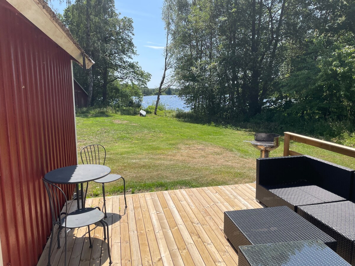 A wooden deck overlooks a grassy area leading to a calm lake. A round table with two chairs is positioned beside a seating arrangement made of dark wicker. The surrounding trees provide a natural backdrop, contributing to a peaceful outdoor environment.