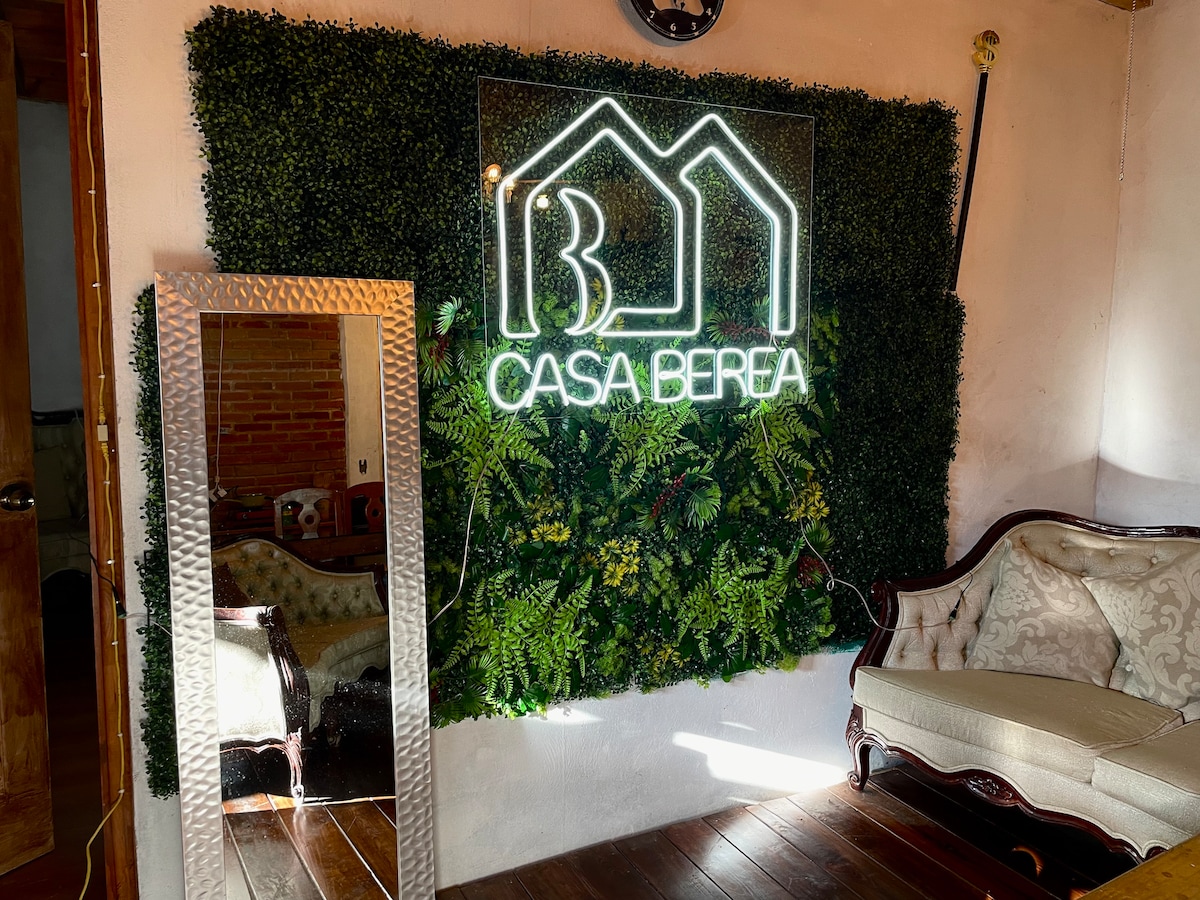 A decorative wall featuring greenery provides a natural backdrop for the illuminated sign that reads 'CASA BERREA.' An elegant vintage sofa is positioned nearby, contributing to the cozy interior atmosphere with rich wooden flooring underfoot.