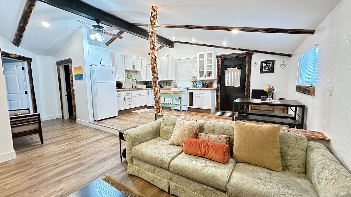 A bright and open living area showcases a comfortable sofa adorned with pillows. A well-equipped kitchen is visible, featuring modern appliances and ample counter space. Natural light filters in through nearby windows, illuminating the inviting space, which seamlessly connects to a dining area.
