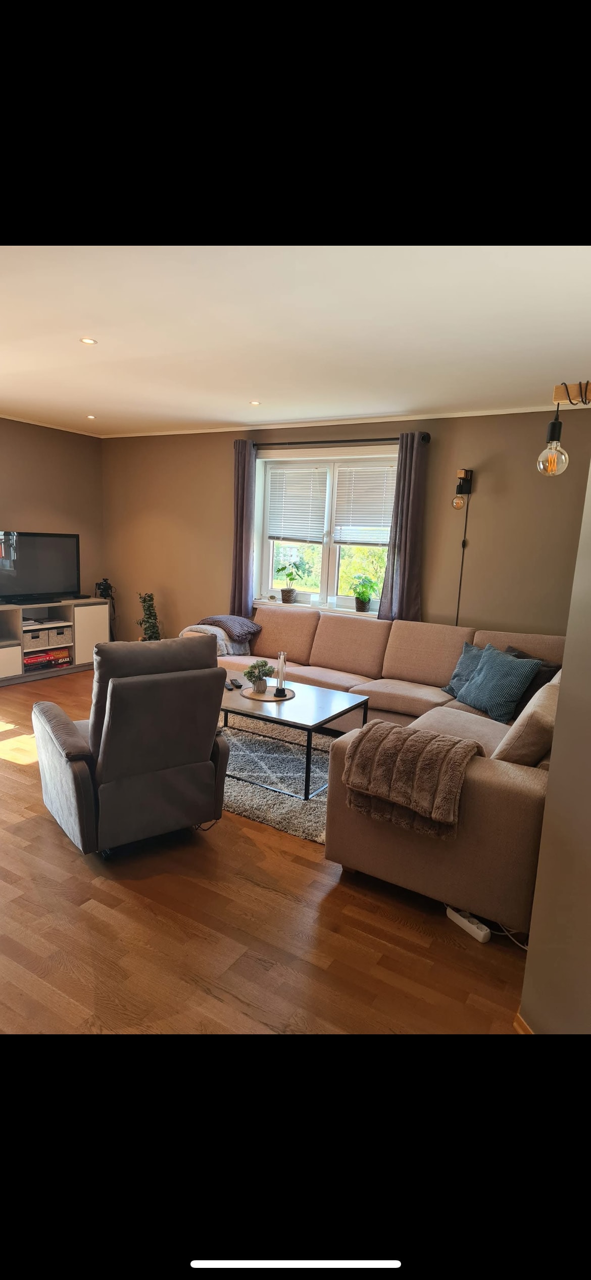 A comfortable living area is furnished with a light-colored sectional sofa and two accent chairs. A modest coffee table sits at the center, with decorative items arranged on top. Natural light filters through a large window dressed with light curtains, illuminating the warm wood flooring.
