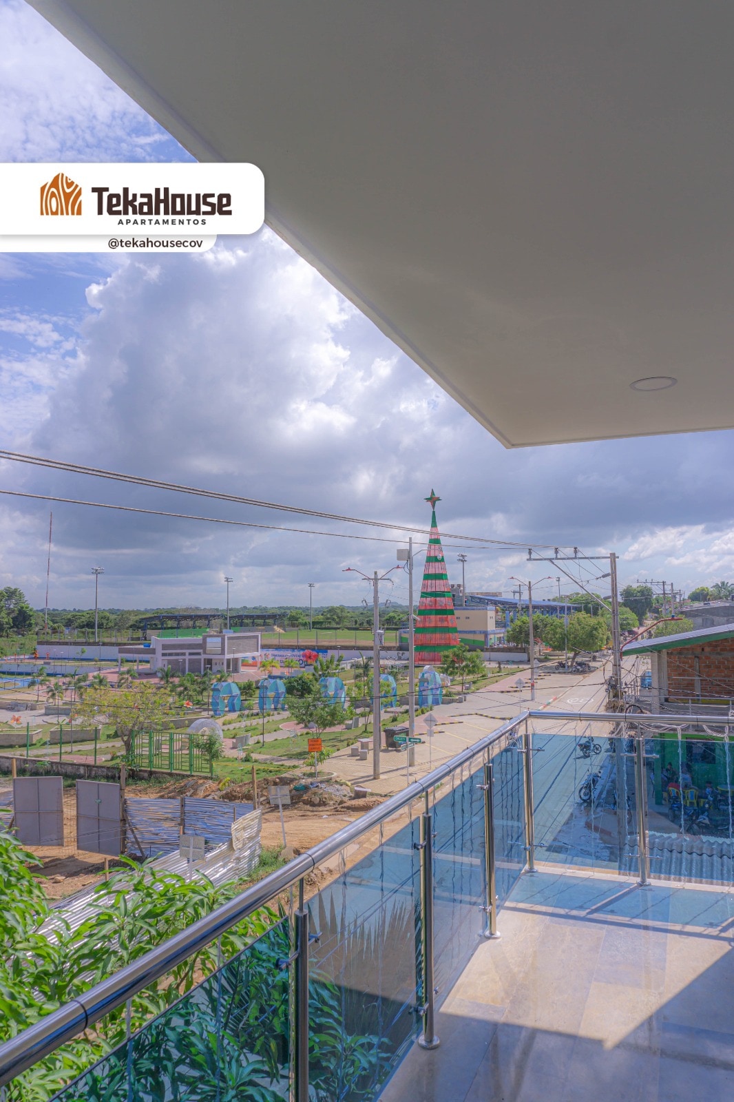 A balcony view reveals a vibrant outdoor area featuring a tall decorative tree in the center, surrounded by greenery and pathways. A railing made of glass offers an unobstructed sight of the landscape, enhanced by a partly cloudy sky.