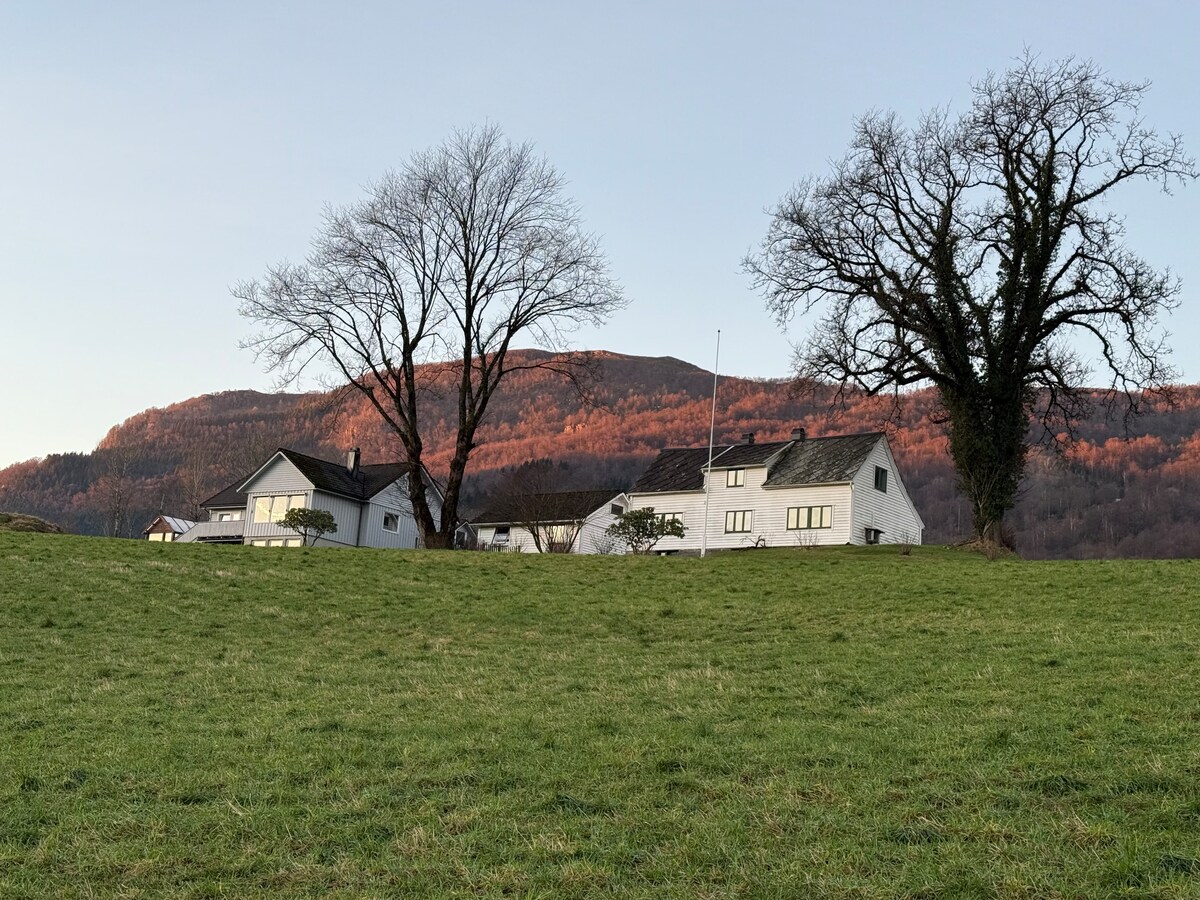 The image depicts a serene farmhouse set against a backdrop of rolling hills. A vibrant landscape features two buildings with large windows and a flagpole, surrounded by open green fields. Leafless trees frame the scene, highlighting the natural beauty of the area.