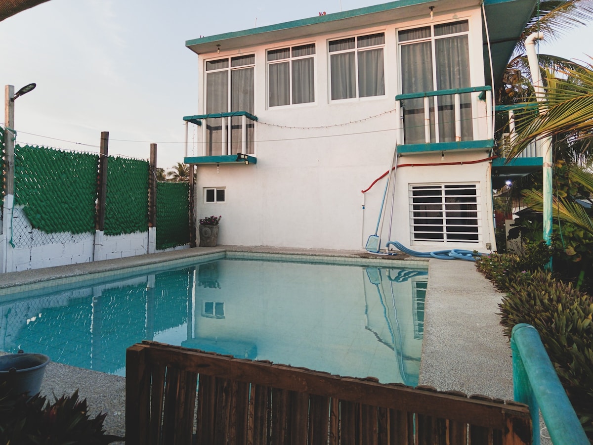 A two-story house with large windows is seen alongside a clear swimming pool. Balconies can be observed on the upper floor, surrounded by foliage. The area is enclosed by a fence, providing privacy for outdoor relaxation near the water.