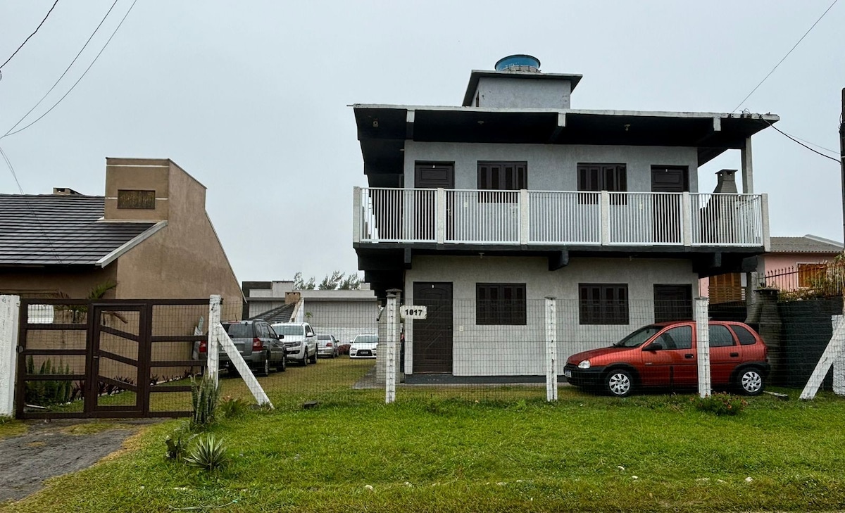 A three-story building is presented, featuring a light grey exterior with dark trim. A balcony with white railings extends from the upper level. The front yard is grassy with a paved driveway and a parked red car. A fence encloses the property, offering a sense of privacy.