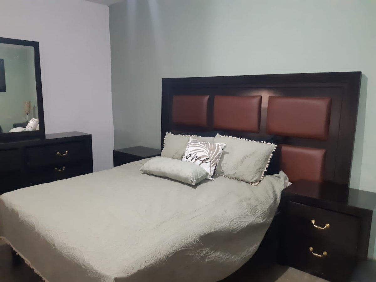 A comfortable sleeping area features a spacious bed with a quilted coverlet and decorative pillows. Dark wood nightstands are placed on either side, complemented by a large mirror above a matching dresser. Soft lighting enhances the soothing atmosphere.