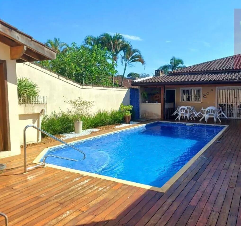 A spacious outdoor area is presented, featuring a swimming pool surrounded by wooden decking. Lush greenery provides a natural backdrop, while patio furniture consisting of white chairs and a table is arranged nearby. A home with an accessible entrance is visible in the background.