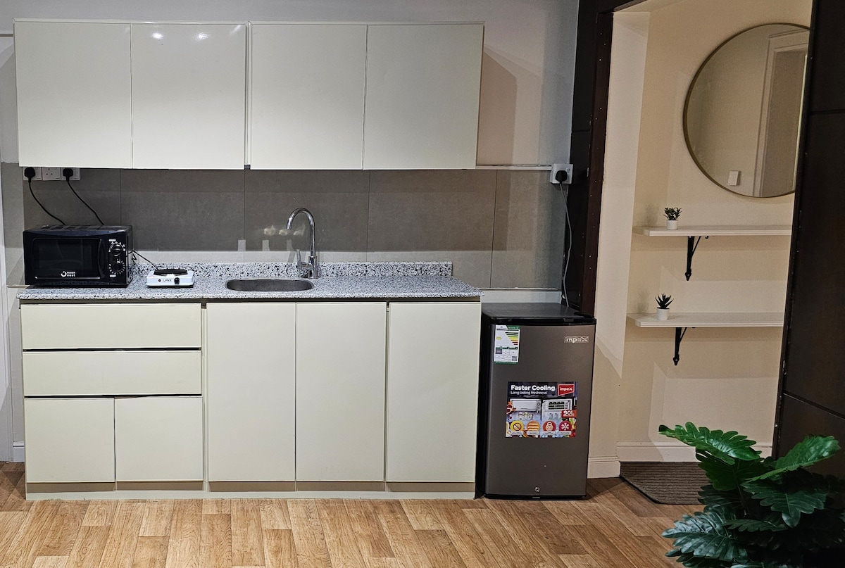 A modern kitchen is designed with sleek, light-colored cabinetry and a countertop featuring a subtle pattern. Essential appliances are visible, including a microwave and a refrigerator. A sink is positioned within easy reach, and a small shelf displays minimal decorative items. Greenery enhances the space.