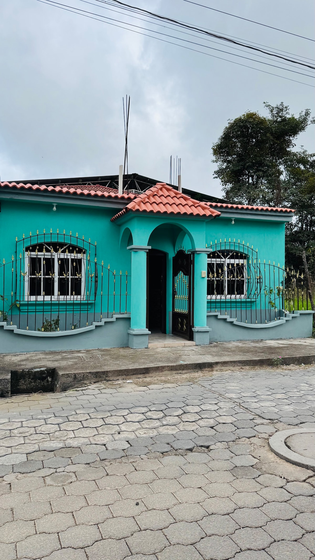 A colorful house with a turquoise exterior is framed by decorative ironwork. The distinctive red tiled roof adds contrast, while multiple windows with arched tops allow natural light to enter. A paved walkway leads up to the entrance, enhancing accessibility.