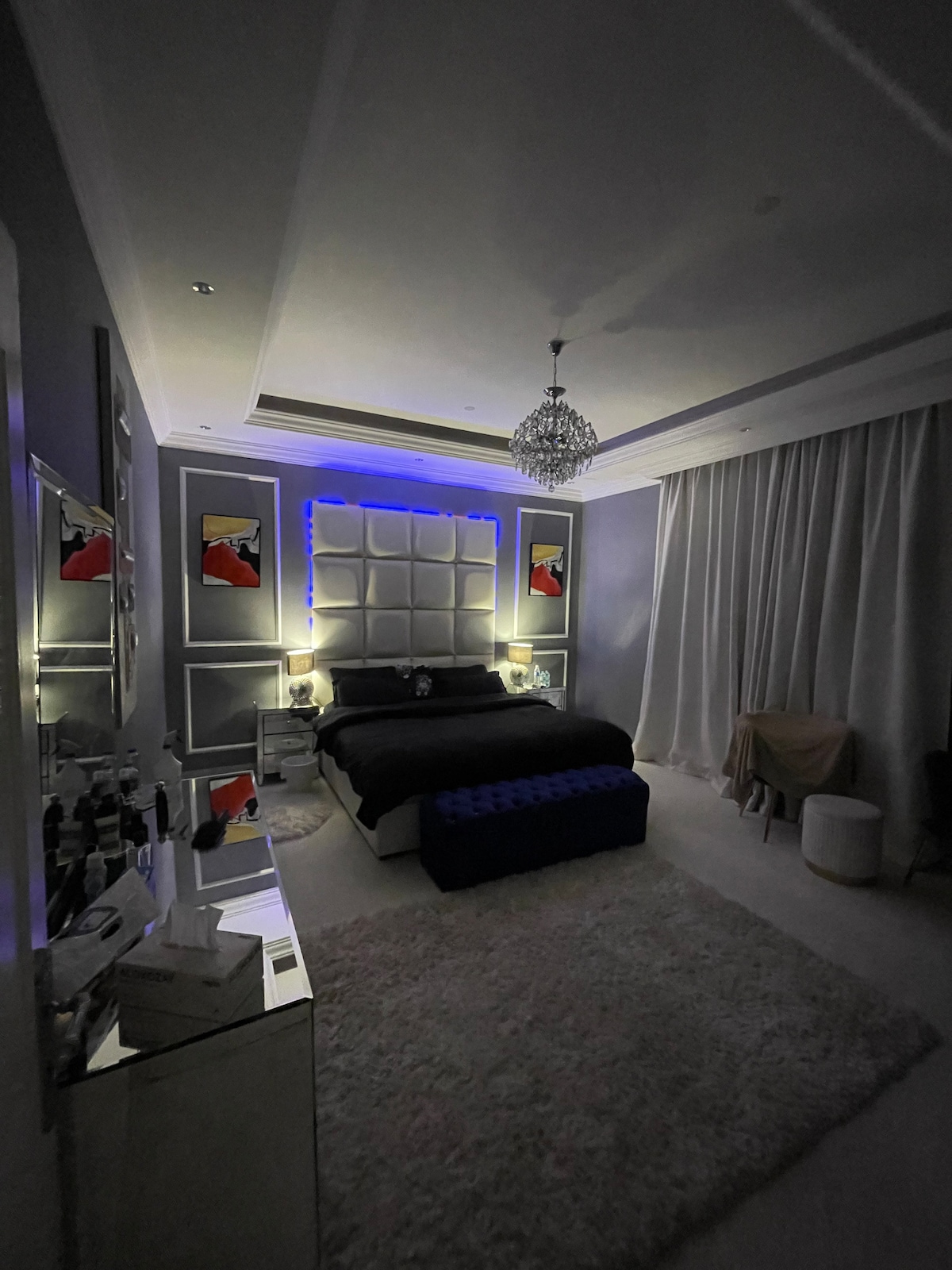 A dimly lit bedroom features a spacious bed adorned with dark bedding. Illuminated artwork hangs on the walls, complemented by a plush area rug on the floor. A chandelier adds elegance, while soft curtains frame a window, creating a serene environment.