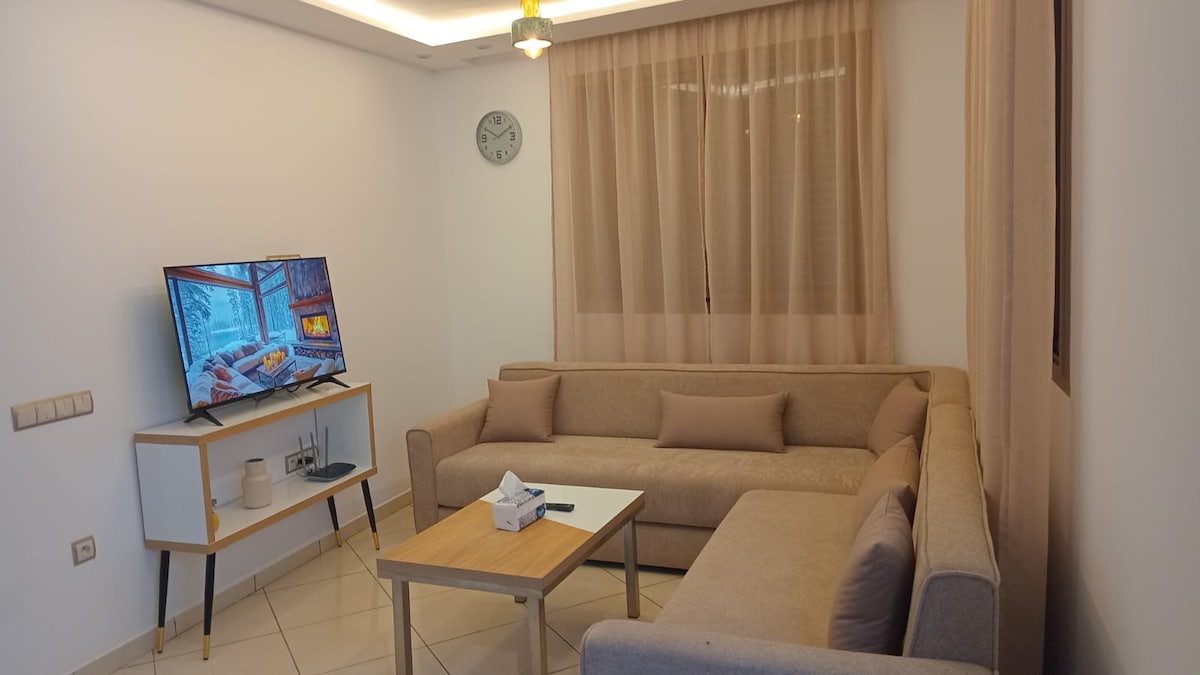 A neatly arranged living area is presented with a light-colored sectional sofa and a minimalist white coffee table. A wall-mounted television is visible next to a round clock, while soft drapes filter natural light through the window.