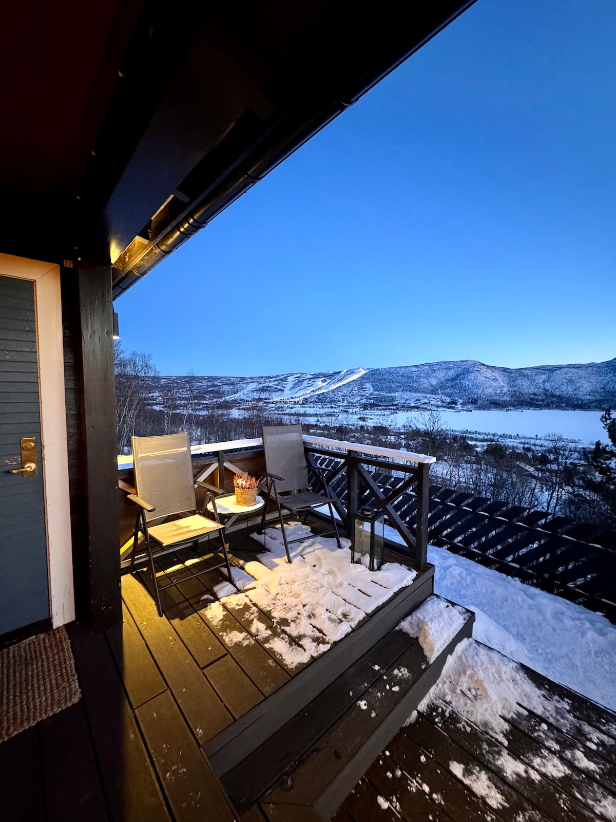 A snowy outdoor terrace features two light grey chairs and a small table, inviting relaxation. The surrounding mountains and valley are visible, creating a scenic backdrop under a clear evening sky. The area is illuminated softly, enhancing the tranquil setting.