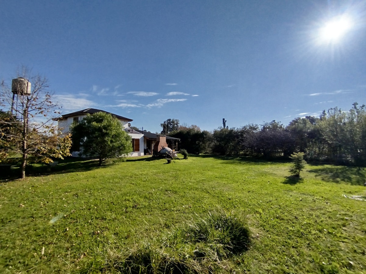 A spacious green lawn is surrounded by trees, creating a tranquil environment. The house is visible in the background, featuring multiple windows and a welcoming façade. A clear blue sky is overhead, with the sun shining brightly, adding to the serene atmosphere.