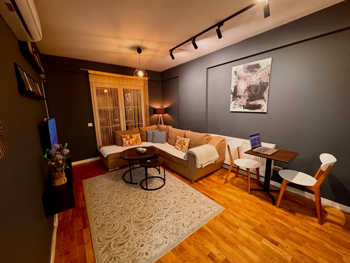 A cozy living room is highlighted by an L-shaped sofa, complemented by a round coffee table. Stylish decor features a large artwork on the wall. Natural light filters through the window, enhancing the warm tones of the wooden floor and furnishings. A compact dining area with two chairs is visible.