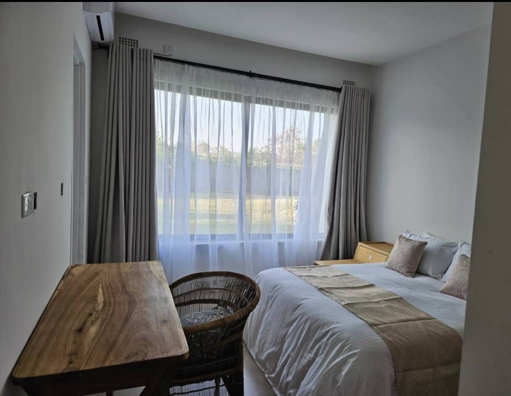 A light-filled bedroom features expansive windows draped with sheer curtains, offering views of the serene garden. A spacious bed is complemented by decorative pillows, while a simple wooden desk and a wicker chair provide a cozy workspace in the corner.