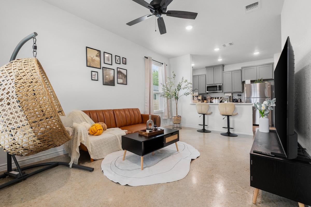 A modern living room features a brown leather sofa with a mustard-colored throw pillow. A round coffee table is centered on a plush area rug. A hanging wicker chair adds texture, while behind, a kitchen with bar stools and stainless-steel appliances is visible. Natural light enters through window treatments.