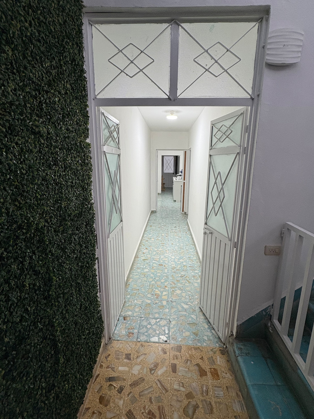 A narrow corridor features a combination of tiled flooring and a textured wall covered in greenery. The doorways at each end provide views of adjacent spaces, with light illuminating the pathway leading into the distance.