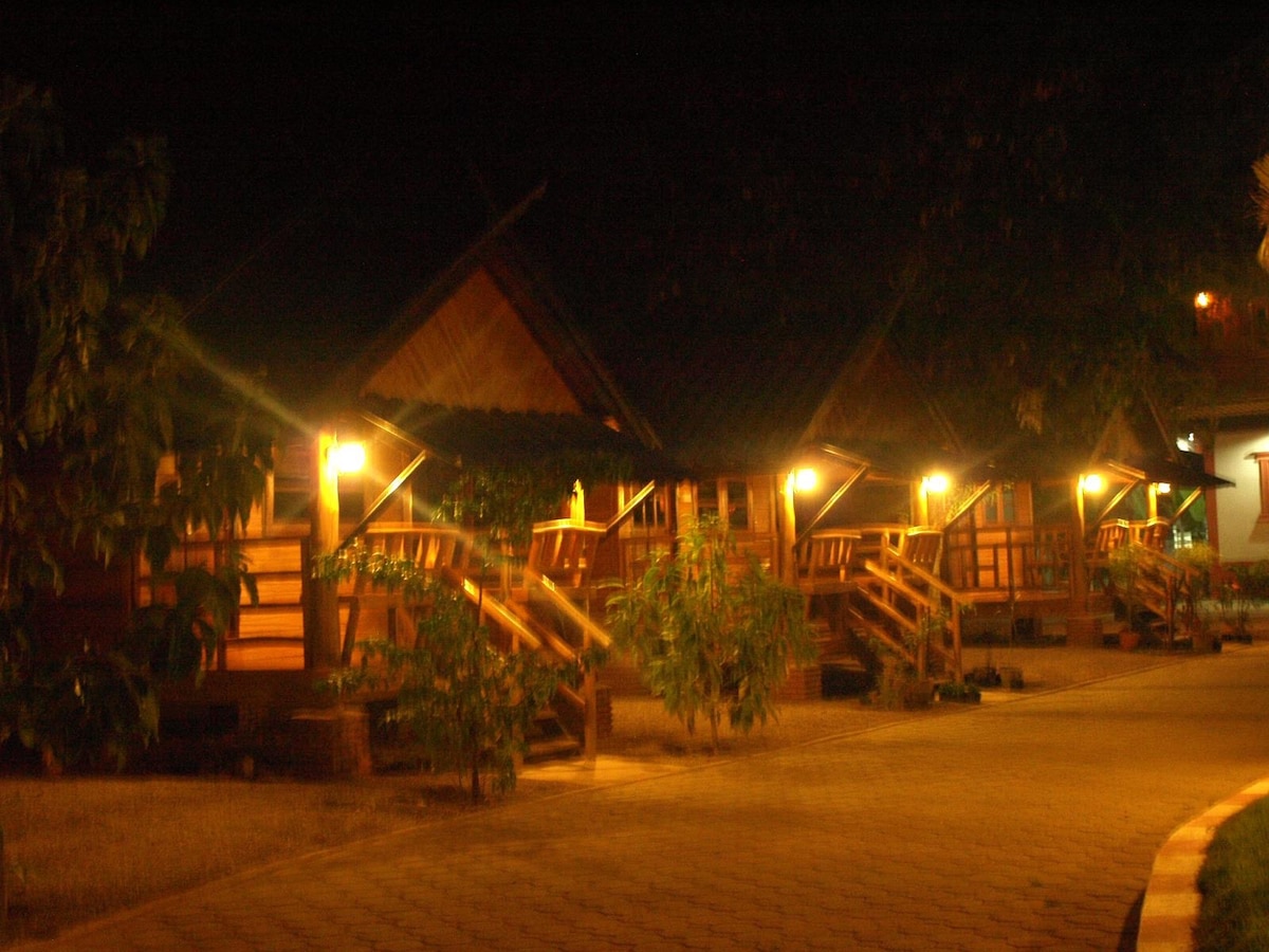 Wooden bungalows are lined along a softly lit pathway, each featuring a porch with steps leading up. Warm light emanates from the bungalows, creating a welcoming ambiance in the evening. Surrounding greenery enhances the natural setting.