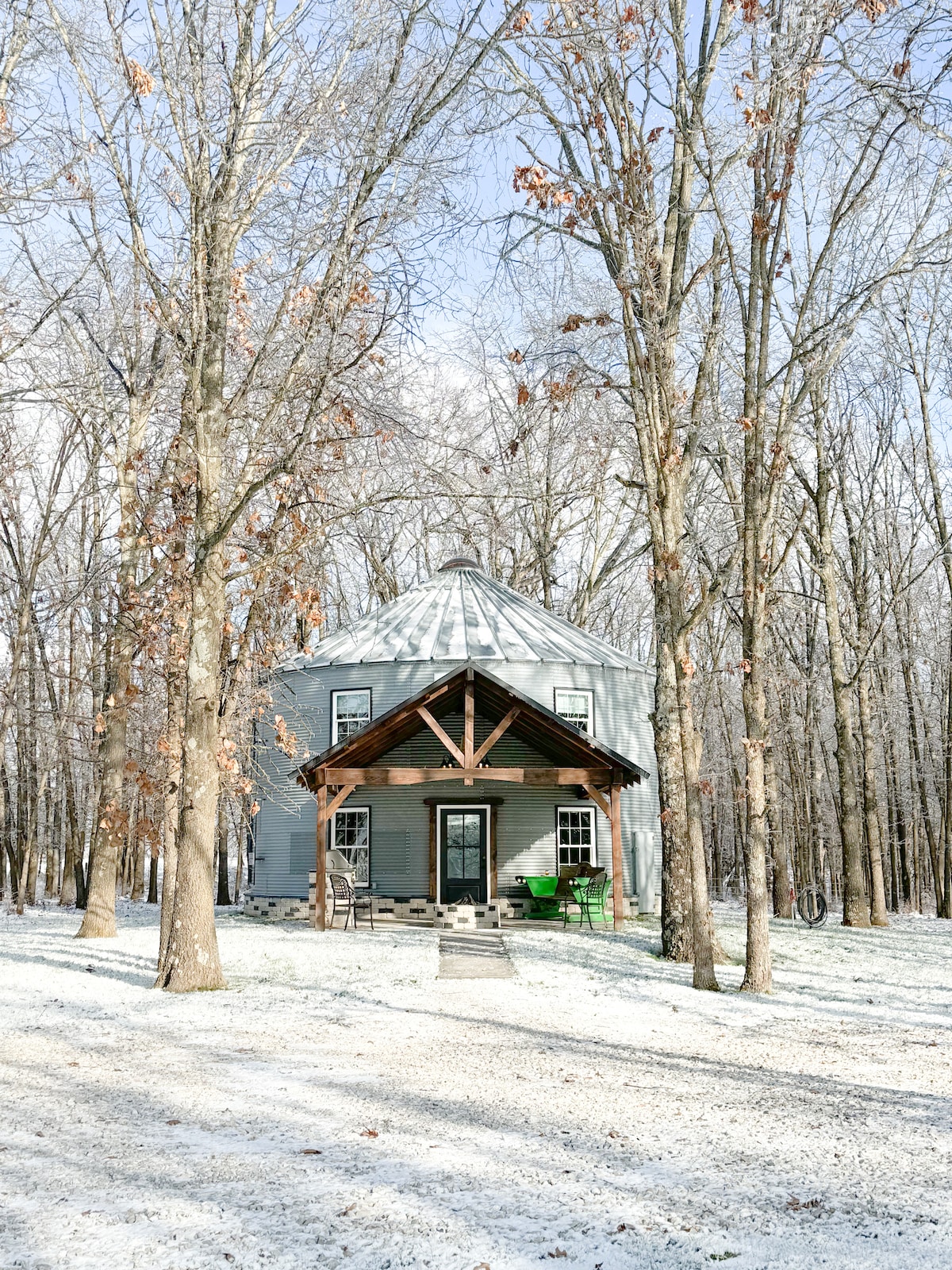 The exterior of a unique grain bin structure is framed by tall trees, with a light dusting of snow covering the ground. A covered porch is supported by wooden beams, and green chairs are placed near the entrance, inviting relaxation amid the serene forest setting.