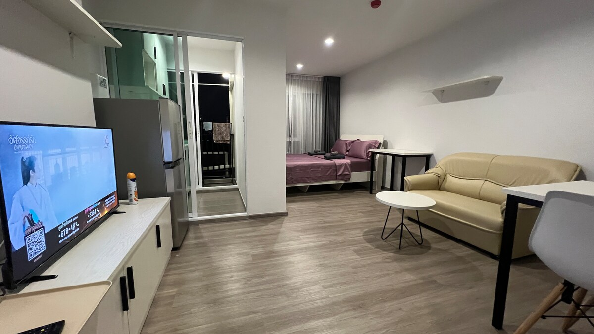 A well-lit living space features a beige sofa and a round coffee table. A mounted television is positioned against the wall. In the background, a spacious bed is visible, complemented by a large window that allows natural light to enter the room.