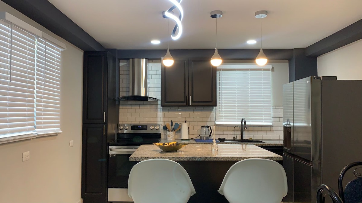 A modern kitchen features granite countertops and dark cabinetry. Three pendant lights illuminate the space, highlighting the tile backsplash. A stainless steel refrigerator stands alongside a stove, while two sleek white chairs are positioned at the counter with a bowl of fruit in the center.