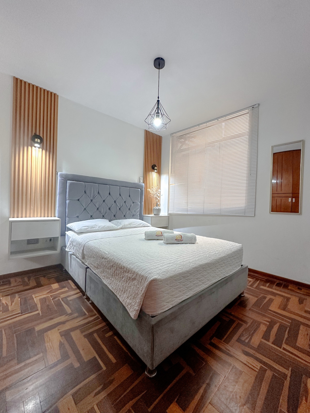 A well-lit bedroom features a spacious bed with a padded headboard and two rolled towels placed at the foot. Natural light filters through the window, and wood accents create a warm atmosphere. Stylish lighting and a mirror add to the room's modern design.