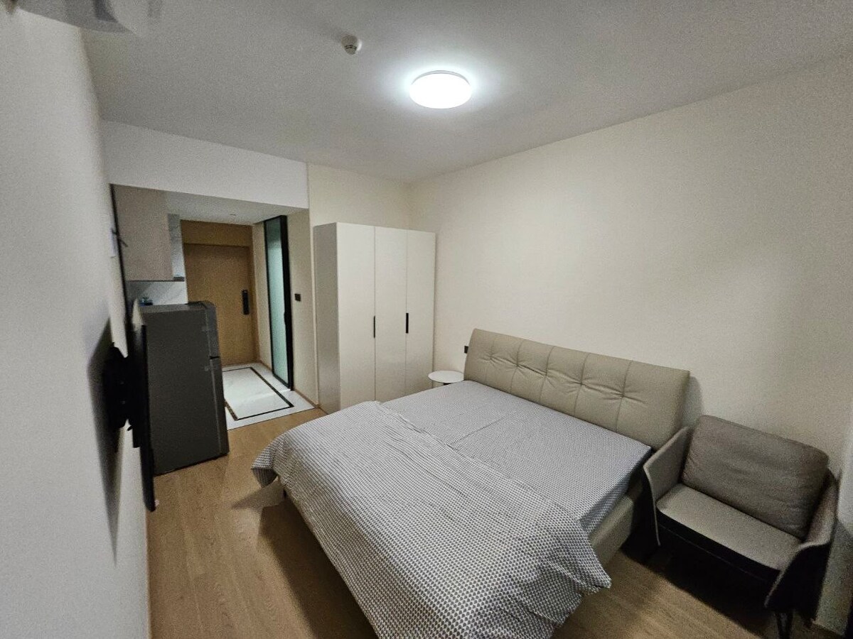 A modern sleeping area features a spacious bed with a quilted headboard and a light-colored coverlet. A small bedside table is positioned next to the bed. Adjacent, a seating area includes a simple chair. Built-in storage provides organization with a clean, minimalist aesthetic.