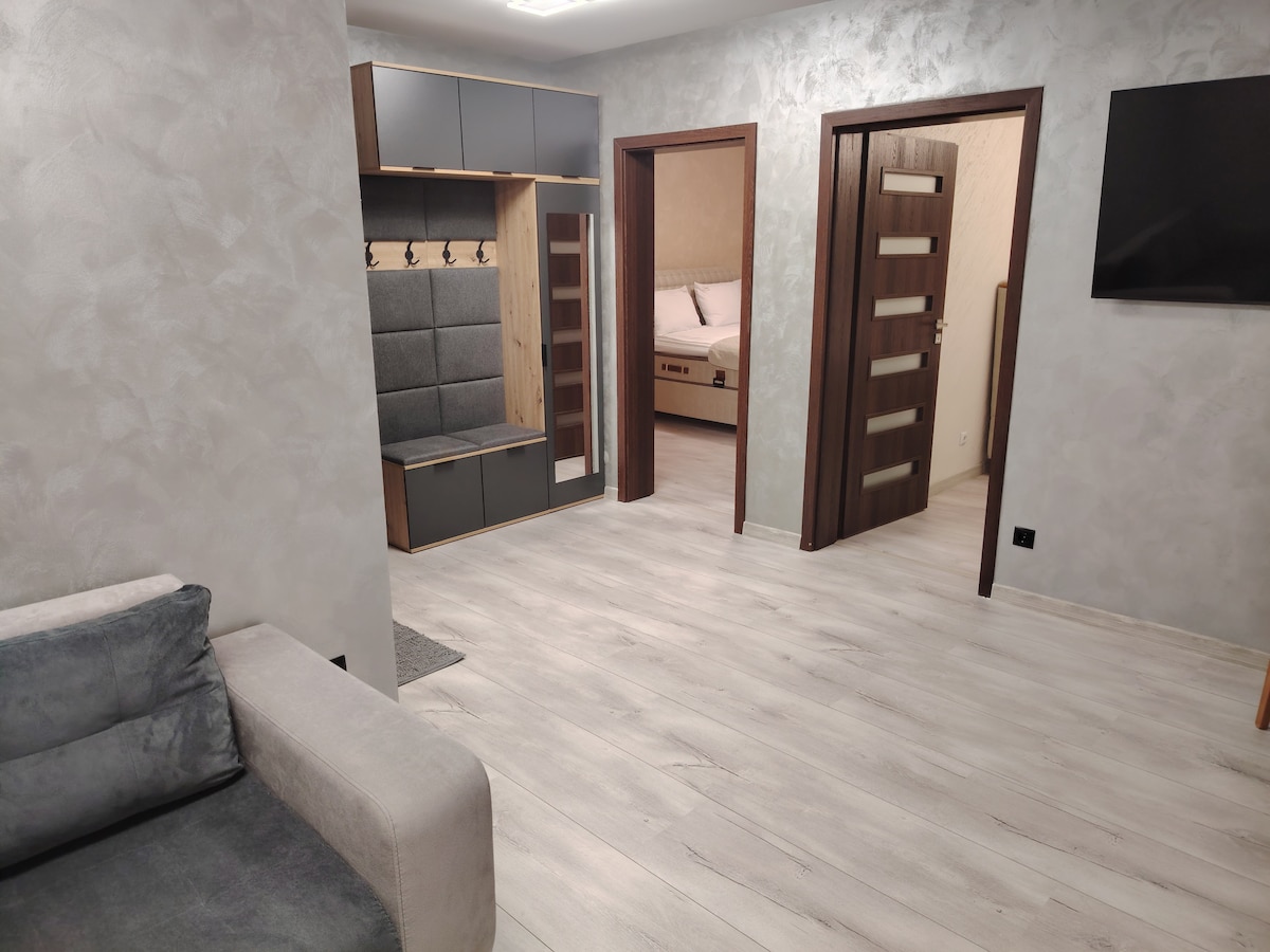 The entrance is highlighted by a modern storage unit featuring hooks and seating. Light-toned wooden flooring complements the neutral wall color. A doorway leads to a bedroom, while a plush couch is positioned against the wall, providing a welcoming touch to the space.
