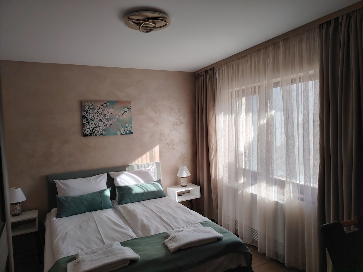 A well-lit bedroom features a spacious bed with crisp white linens and contrasting green accents. Two bedside tables with lamps are positioned on either side. A large window, draped with sheer curtains, allows natural light to filter in, enhancing the soft tones of the room.