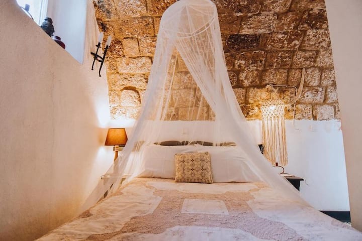 A cozy sleeping area features a spacious bed draped with a delicate, sheer canopy. The room showcases textured stone walls and warm lighting from a nearby lamp, creating a serene environment. A decorative pillow adds a subtle touch to the bed's inviting presentation.