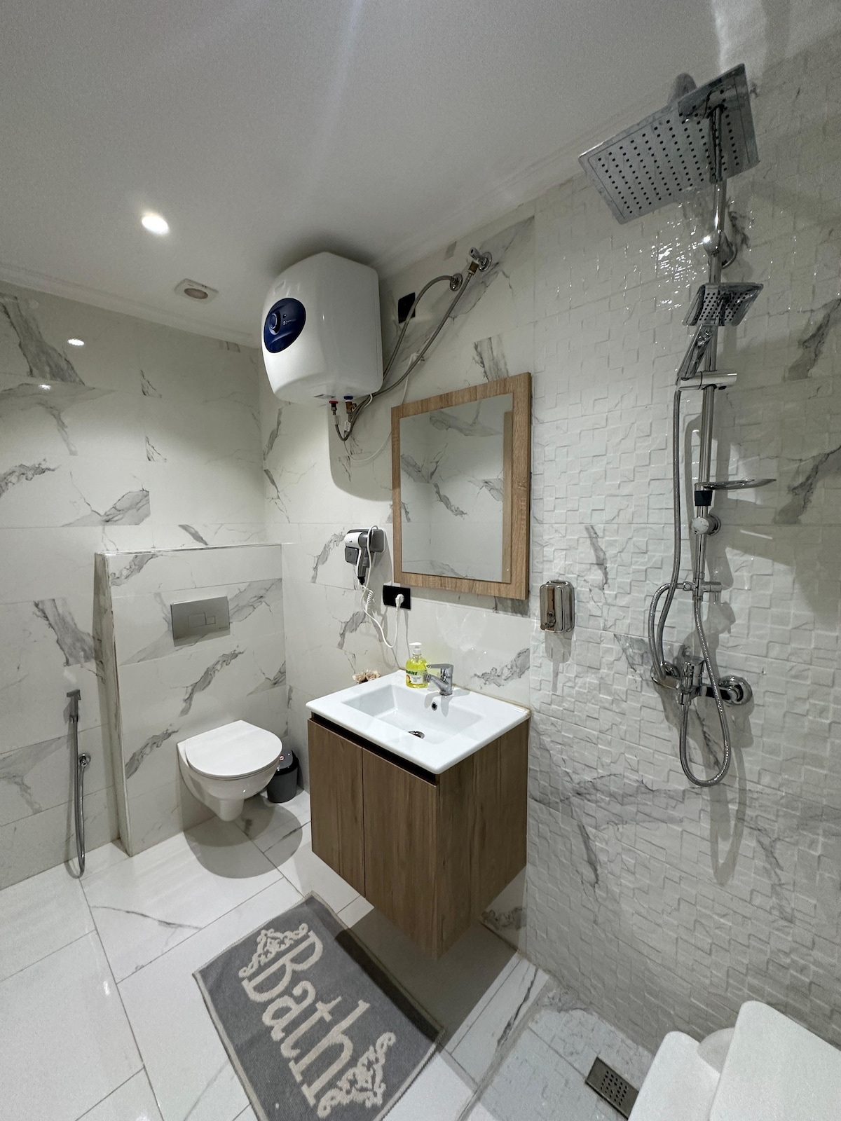 A modern bathroom is presented with white marble-patterned walls. A compact vanity with a wooden finish features a sink and a mirror above. A shower with a glass partition is visible, along with a toilet positioned discreetly. A soft rug displaying the word 'Bath' lies on the floor.