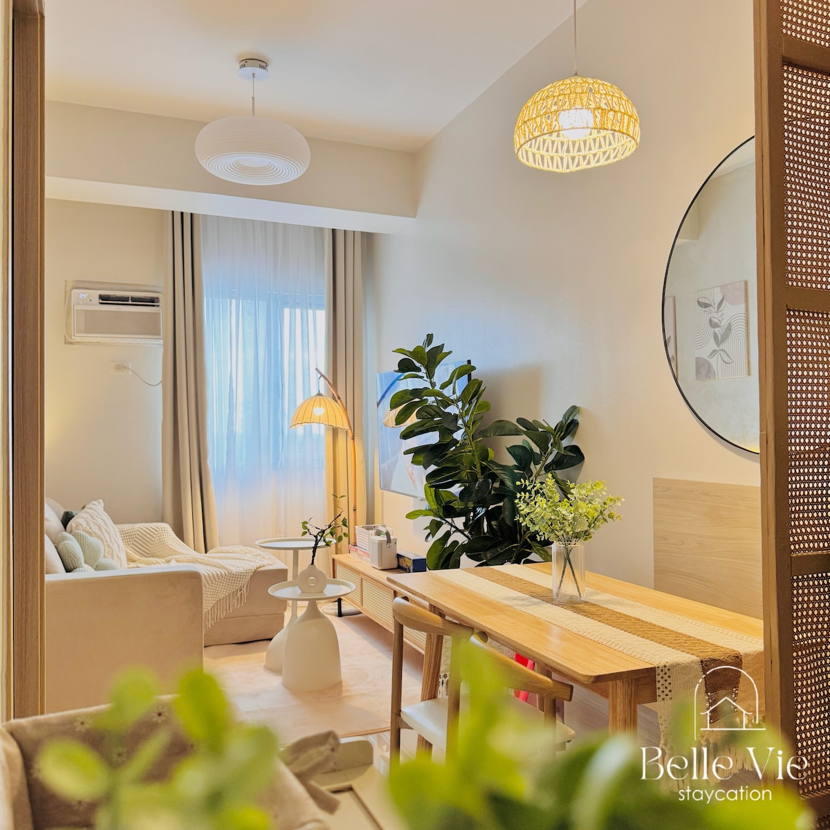 A well-lit living area features a comfortable L-type sofa, a stylish floor lamp, and a dining table adorned with a decorative centerpiece. Natural light filters through sheer curtains, creating a serene environment complemented by potted plants and minimalist decor.