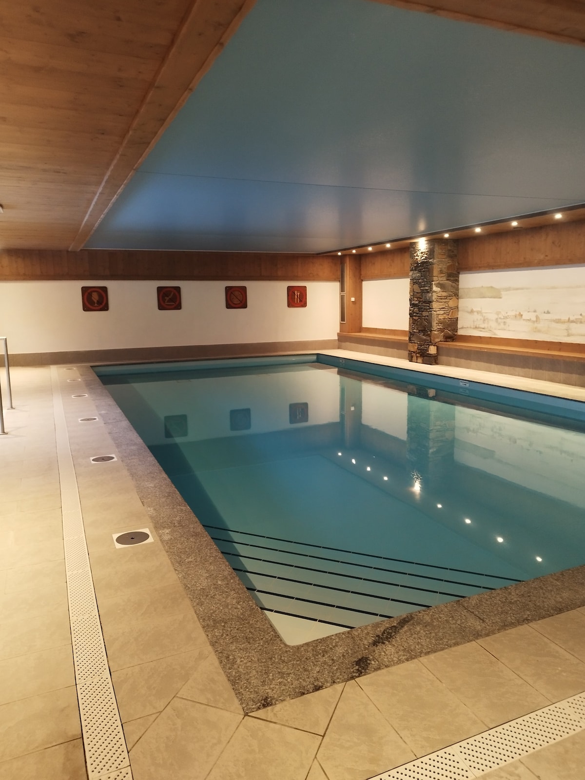 An indoor swimming pool is featured, surrounded by neutral-toned walls and illuminated by soft lighting. The water appears calm and inviting, reflecting the blue ceiling above. Textured stone columns and decorative wall panels add subtle interest to the serene environment.